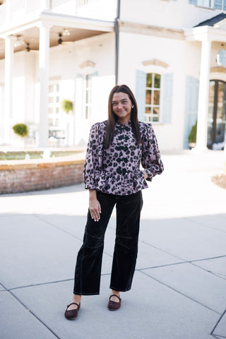 Woman posing outdoors in a mauve and black leopard print peplum blouse featuring dramatic puff three-quarter sleeves, a mandarin collar, and decorative black frog closure knots down the front. The blouse is paired with black velvet contrast-stitched trousers and dark brown Mary Jane flats against a bright, modern courtyard background.