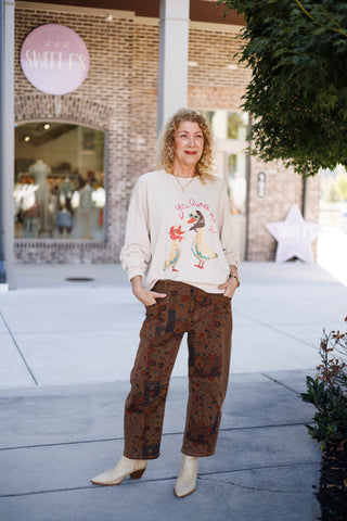 Woman smiling and posing outdoors wearing high-waisted dark brown utility trousers featuring vintage floral tapestry patchwork panels down the legs in rich autumn hues of rust, navy, and mustard. Styled casually with a cream graphic sweatshirt, pointed-toe cream snakeskin western boots, and subtle gold jewelry in front of a storefront.