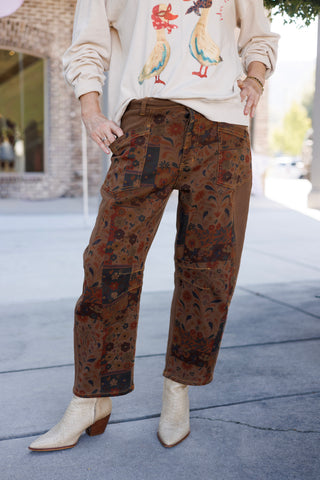 A detailed shot focusing on the lower body showcasing the relaxed, straight-leg cut of the brown utility pants. Highlights the front floral tapestry paneling, mustard contrast stitching, large utility patch pockets, and button fly, paired with cream snakeskin ankle boots.