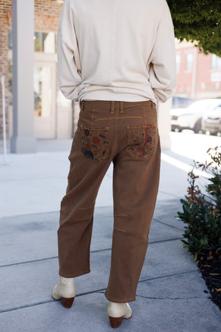 Back view from the waist down highlighting the solid dark brown canvas construction of the trousers. Features twin back patch pockets lined with matching floral tapestry print, mustard topstitching along the waist and seams, knee-seam detailing, and a comfortable relaxed silhouette
