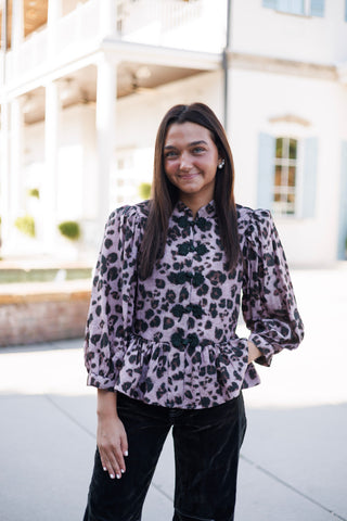 Three-quarter portrait of a smiling woman wearing a mauve pink leopard-patterned peplum top with black Asian-inspired frog closure ties along the front closure. Styled with one hand in her pocket, showcasing the gathered flared peplum hem and structured puff shoulders paired with dark trousers.