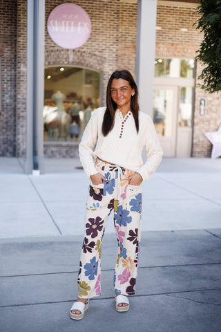 Woman posing outdoors in a cozy cream-colored cable-knit Henley sweater featuring a split collar placket lined with tortoise-shell buttons. The long-sleeve sweater is paired with wide-leg cream sweatpants covered in a colorful retro floral print, detailed with a white contrast drawstring waist and front patch pockets. Styled with white platform slide sandals in front of a brick storefront.
