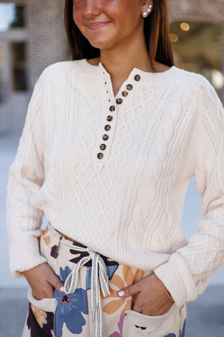 Front-facing mid-shot showcasing the cable-knit texturing and buttoned placket of a cream Henley pullover sweater. The sweater is worn casually over the drawstring waistband of multicolor floral lounge trousers, accessorized with pearl stud earrings.