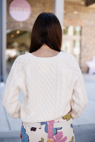 Back view of a woman wearing a cream cable-knit sweater, displaying the continuous diamond and cable stitching across the back panel and raglan-style sleeve construction. Styled resting right above the high waistband of multicolor floral pants.