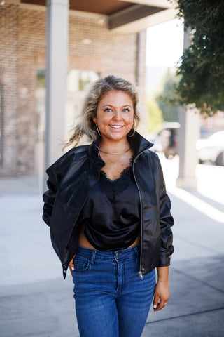 Woman posing outdoors wearing an oversized black faux-leather bomber jacket styled open over a black satin lace-trim camisole top and classic blue denim jeans. She stands smiling with one hand in her pocket, accessorized with gold hoop earrings and a minimal choker necklace against a modern storefront backdrop.