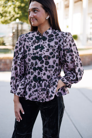 Close-up portrait highlighting the mandarin standing collar, volume-puffed sleeve shoulders, and intricate black floral frog closure knots of a dusty mauve leopard printed top. Styled with classic pearl stud earrings and dark velvet pants.   