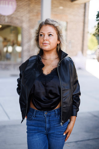 Mid-length front view of a woman wearing a black zip-front faux-leather jacket featuring a stand collar, dropped shoulders, and silver zipper hardware. The jacket is layered open over a black lace-trim silk cami and high-waisted medium-wash denim jeans.