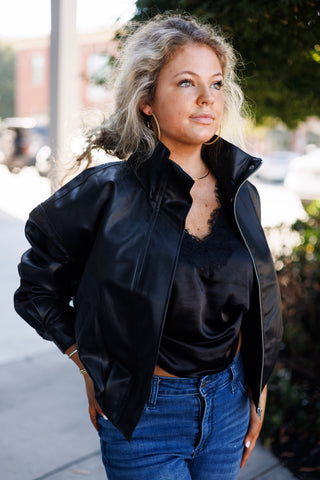 Three-quarter angled shot highlighting a black faux-leather bomber jacket with a relaxed silhouette and structured collar details. Worn open to reveal a black lace-trimmed satin top tucked into blue jeans, styled with gold hoop earrings and delicate jewelry.