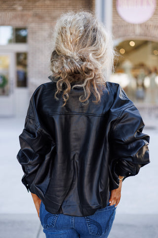Back detail shot of a woman wearing a relaxed-fit black faux-leather bomber jacket showing the clean horizontal back yoke stitching and wide slouchy sleeve construction. She wears her curly blonde hair in a casual updo above high-waisted blue jeans.
