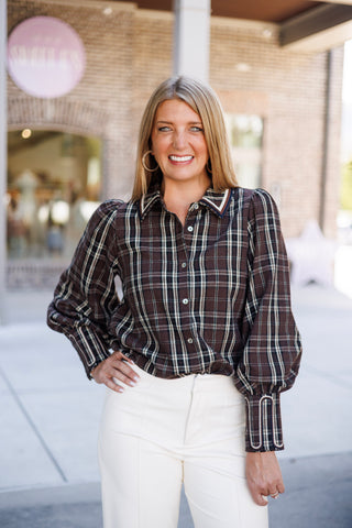 Woman smiling outdoors posing in a chocolate brown plaid long-sleeve blouse featuring white, tan, and black tartan grid lines. The blouse features a crisp point collar bordered with multi-colored striped trim, button-front closure, voluminous puff sleeves, and wide extended cuffs with stitched embroidery detailing, styled tucked into off-white tailored trousers
