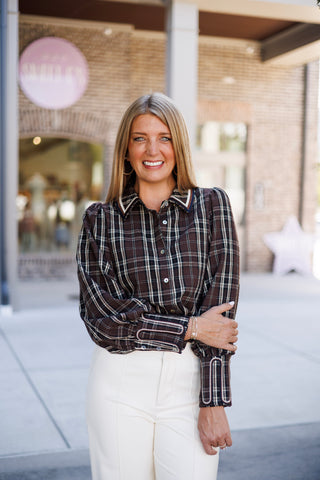 Upper body shot highlighting the tailored structure of the brown plaid statement blouse. Showcases the point collar with contrast navy and cream edging, mother-of-pearl front buttons, and elongated barrel cuffs with decorative arched stitching, paired with high-waisted white pants with center seam detail.   