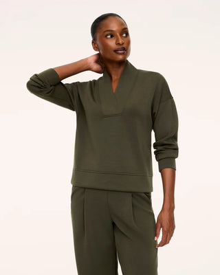 Front three-quarter view of a model in the Monstera olive green AirEssentials pullover sweatshirt paired with matching pleated track pants. The design highlights an elegant folded overlap V-neck collar, lightweight spacer fabric with an ultra-soft matte finish, push-up ribbed sleeve cuffs, and a straight waistband hitting right at the hips.