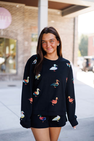 Woman smiling outdoors in front of a brick storefront, wearing a black two-piece printed fleece lounge set featuring an all-over colorful watercolor duck motif. The outfit includes an relaxed crewneck long-sleeve sweatshirt with ribbed trim paired with matching high-waisted elastic lounge shorts.