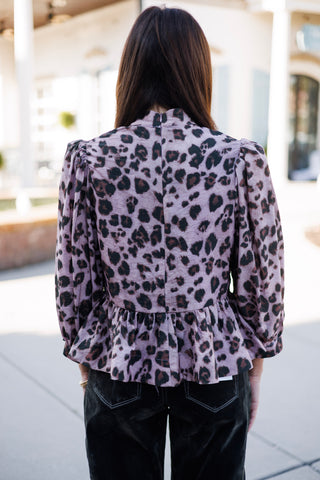 Back view of a woman wearing a dusty mauve leopard spot peplum blouse showing the high standing collar with a subtle zip closure and the gathered, flared peplum waist seam. The view highlights the structured volume of the puff sleeves and dark contrast-stitch back pockets on her trousers.