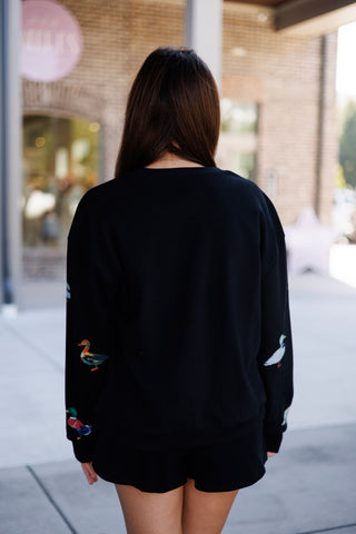 Full back view of the black printed lounge set showing the plain back of the crewneck sweatshirt paired with matching black fleece shorts that feature subtle duck prints along the sides and hem. Styled with straight brown hair worn loose