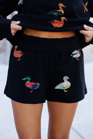 Detailed close-up shot of the high-waisted black fleece lounge shorts showing the thick elastic waistband and front duck illustrations, with the hem of the matching printed crewneck sweater lifted slightly at the sides.