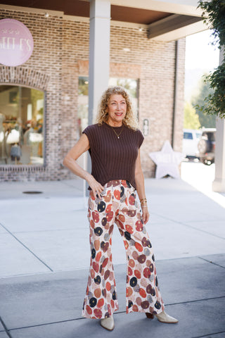 Woman posing in a sleeveless chocolate brown ribbed knit vest featuring a round crew neckline and architectural chevron ribbing at the hem. The sweater vest is paired with high-waisted wide-leg trousers featuring a bold abstract geometric print in rust red, black, beige, and gold foil accents. Styled with cream-colored pointed-toe boots and dainty gold jewelry against an urban storefront setting.
