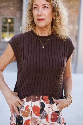 A portrait view showcasing the rich knit texture and ribbed construction of a sleeveless dark brown sweater top with cap-sleeve coverage. Accentuated by a delicate gold paperclip chain necklace with a solitaire pendant, showing hands tucked into the front pockets of high-waisted printed pants.