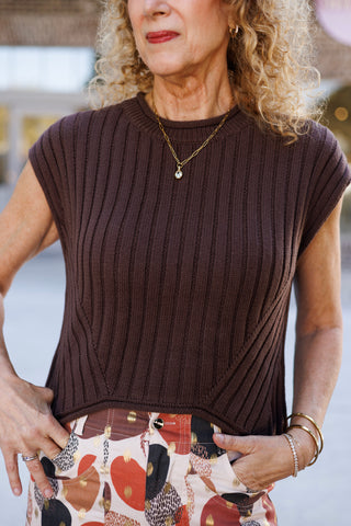 Close-up shot focusing on the high crew neck, fine ribbing, and subtle dropped shoulder design of a sleeveless brown knit vest. The view highlights the top button detail and metallic gold foil dots on the waistband of the colorful patterned trousers.