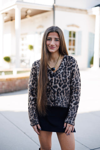 Woman smiling outdoors on a paved walkway in a fuzzy, plush button-up cardigan sweater featuring an all-over leopard animal print in tan, beige, and black tones. The top features a subtle V-neckline, dark tortoiseshell button closures, long sleeves, and a relaxed cropped hemline, styled over a black mini skirt with a statement gold beaded necklace featuring a large black stone pendant.
