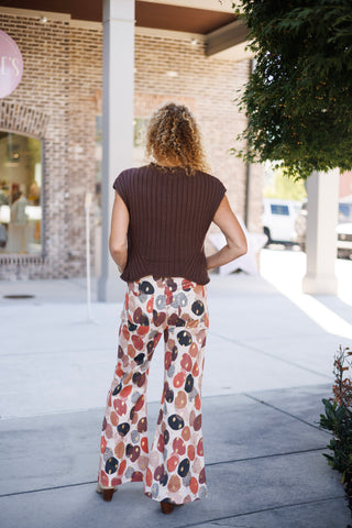 Back view of the outfit showing the straight-back hem design of the dark brown ribbed knit vest and the flowing wide-leg fit of the abstract geometric printed pants. The model stands with hands resting on her hips, showcasing the full flare leg silhouette and subtle heel of her neutral boots.