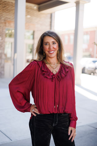 Three-quarter portrait of a smiling woman wearing a rich wine red satin blouse designed with dramatic ruffled collar trim along the neckline and adjustable tassel ties. Posed with one hand on her hip, showcasing the subtle crinkle-satin fabric texture and elegant gathered cuffs paired with black contrast-stitch denim.