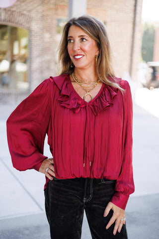 Woman posing outdoors in a luxurious deep burgundy satin long-sleeve blouse featuring a cascading ruffled collar, V-neckline with self-tie drawstring aglets, and voluminous bishop sleeves. The top is styled blouson-tucked into high-waisted black trousers with white contrast stitching, accessorized with layered gold chain necklaces and an oval ring pendant against a paved walkway backdrop