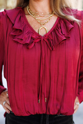 Close-up shot highlighting the detailed V-neckline, layered ruffle collar, and metal-tipped front tie details of a deep red satin blouse. The rich texture of the gathered satin fabric is complemented by stacked gold link chains and a prominent open-oval pendant.