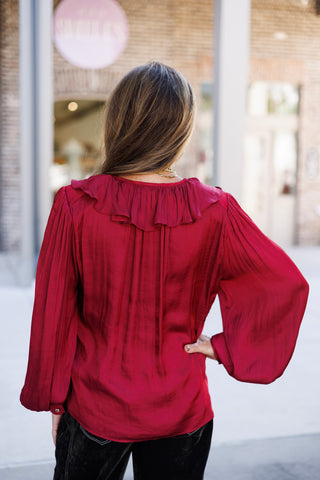 Back view of a woman wearing a rich burgundy satin blouse showing the wide ruffle collar extending across the back neckline and fine vertical pleating along the back yoke. The view highlights the long, fluid bishop sleeves with buttoned cuffs paired with dark pants.