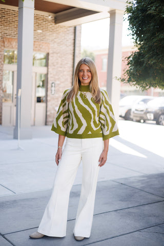 Woman smiling and standing outdoors wearing a relaxed olive green and cream zebra-pattern knit sweater featuring wide three-quarter sleeves, a modest mock neck, and ribbed trim. Paired with high-waisted white wide-leg trousers, pointed-toe textured flats, a chunky gold ring pendant necklace, and bracelets in front of an upscale brick storefront.