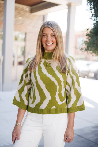 Portrait view of the olive green and cream zebra print sweater showcasing the fine-knit texture, soft mock neck detail, and oversized gold ring pendant necklace. Shows a close look at the boxy three-quarter sleeves and warm neutral colorway.