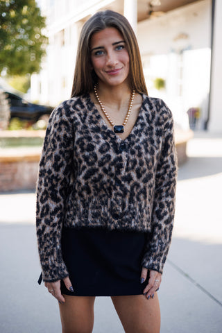 Close-up portrait view emphasizing the plush mohair-like texture and detailed spot pattern of the leopard sweater. Displays the deep V-neck cut, dark button closures, and fine fuzzy knit finish, accessorized with a gold beaded necklace featuring an emerald-cut black gem.
