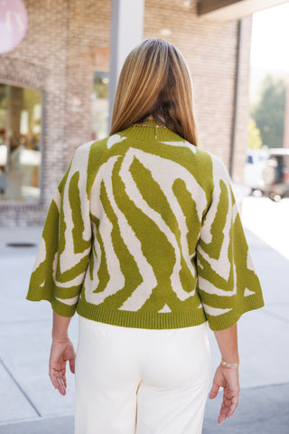 Back view of the olive green and cream zebra jacquard sweater highlighting the seamless continuation of the bold animal print across the back and wide elbow-length sleeves. Styled with white tailored trousers and blonde hair falling past the shoulders over a gold link chain.