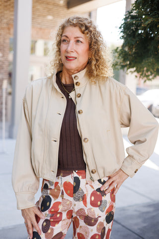 Woman with blonde curly hair posing outdoors wearing a cream canvas button-front bomber jacket featuring a high stand collar, tortoiseshell button front closure over a full zipper, side welt pockets, drop shoulders, and an elasticated hem. Layered over a dark brown ribbed top and paired with high-waisted printed trousers with metallic gold accents