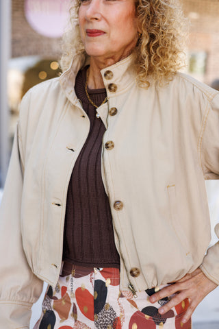 Detailed close-up portrait of the cream utility bomber jacket showing the stand collar with button tab, front zipper closure with tortoiseshell accent buttons, gold topstitching, and subtle drop shoulder seam design layered over a brown knit top.