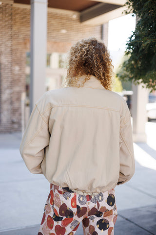Back view from the waist up of the cream casual bomber jacket showing the clean back yoke detail with yellow contrast stitching, relaxed fit, gather at the elastic hemline, and relaxed long sleeves.   
