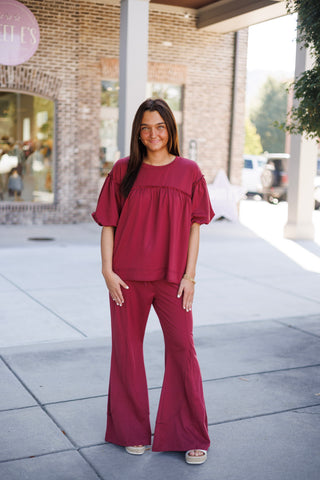 Woman smiling outdoors wearing a rich crimson red two-piece lounge set comprising a short-sleeve puff-sleeve top with gathered chest detail and high-waisted wide-leg flare lounge pants. Styled casually with white platform espadrille slide sandals in front of a modern brick storefront.   