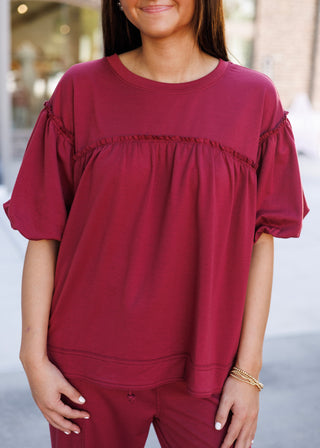 A portrait view of the Crimson Puff-Sleeve Loungewear Top highlighting the delicate ruffle trim that accents the chest yoke and shoulder seams. Crafted from a soft, rich burgundy-red cotton knit, this top offers an elevated casual look with gathered empire seam detailing and comfortable elasticated puff cuffs.