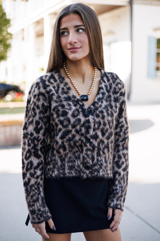 Mid-length front portrait highlighting the soft, textured knit fabric of the leopard print cardigan. Showcases the dark button placket, cozy long sleeves, relaxed silhouette, and V-neck cut paired with black bottoms and gold statement jewelry set against an outdoor building courtyard.
