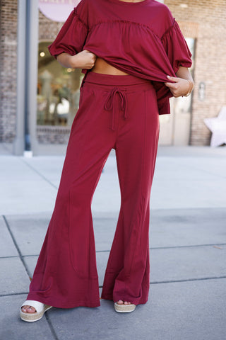 A detailed shot focusing on the lower body showcasing the flare lounge pants, highlighting the thick ribbed elastic waistband with a matching drawstring, center leg seam lines, and wide bell-bottom hems. The top hem is pulled up slightly to show the high-rise waistband fit and comfortable cotton-blend knit fabric