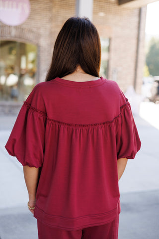 Back view of the Crimson Puff-Sleeve Loungewear Top showcasing the continuous horizontal ruffle trim seam across the upper back and shoulders. The relaxed silhouette flows down to a wide, double-stitched hem, highlighting the soft drape and breathable fit of the elbow-length puff sleeves.