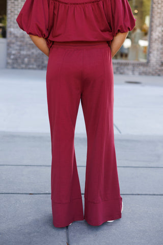 Back view from the waist down displaying the smooth, contoured fit and flare silhouette of the burgundy-crimson knit lounge pants. Highlights the clean back waistband, vertical center leg seams, wide hem cuffing, and the gathered lower hem of the matching short-sleeve top.