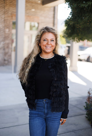 Woman smiling outdoors wearing an open-front black textured faux-fur fringe vest layered over a black long-sleeve ribbed sweater top with subtle white trim at the crew collar and cuffs. Styled with high-waisted blue jeans, gold hoop earrings, and a delicate gold script necklace in front of a brick storefront.