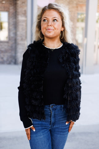 Mid-length front view showcasing a sleeveless black shaggy faux-fur vest styled open over a fine ribbed knit long-sleeve sweater. The vest features layered tiers of textured fringe, paired with medium-wash blue denim jeans and gold statement jewelry.