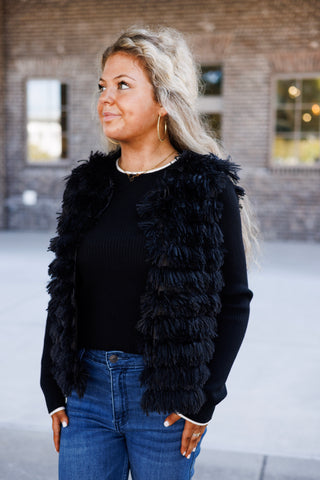 Three-quarter portrait of a woman looking to the side in a black fringe faux-fur vest showing the dimensional texture and open-front layering design. Worn over a fitted black long-sleeve top and blue denim trousers.