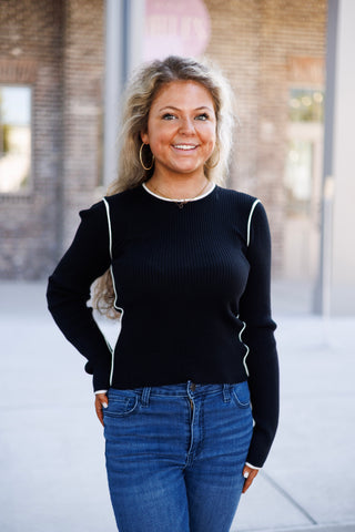 Woman smiling outdoors wearing a fitted black long-sleeve ribbed knit top featuring thin off-white contrast piping along the crew neckline, armhole seams, and side bodice seams. Styled casually with high-waisted medium-wash blue denim jeans, gold hoop earrings, and a delicate gold script necklace