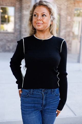 Three-quarter portrait of a woman posing in a black ribbed knit long-sleeve sweater top with cream contrast trim highlighting the shoulder armhole seams, side contours, and sleeve cuffs. Paired with classic blue jeans and minimalist gold jewelry against a modern brick background.