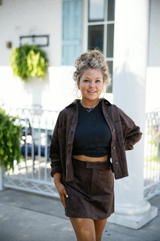 Woman posing outdoors in a chocolate brown faux suede two-piece set, featuring a cropped button-down shacket over a black fitted crop top and a matching high-waisted faux suede mini skirt. Styled with gold hoop earrings, a delicate script necklace, and an updo hairstyle in front of a white building background.