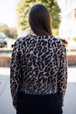 Back view from the waist up of the fuzzy leopard print knit cardigan, showing the continuous animal print pattern across the back and long sleeves, long straight brown hair worn loose, and a straight cropped hemline.