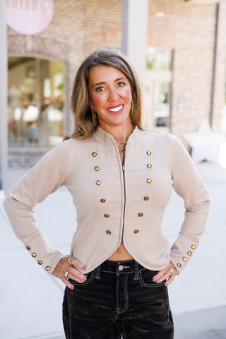 Three-quarter portrait of a smiling woman wearing a beige long-sleeve zip-up sweater jacket with napolean-inspired brass button embellishments. Styled with one hand resting at her hip, showing off the curved front hem cut and decorative sleeve cuffs paired with black contrast-stitch denim.
